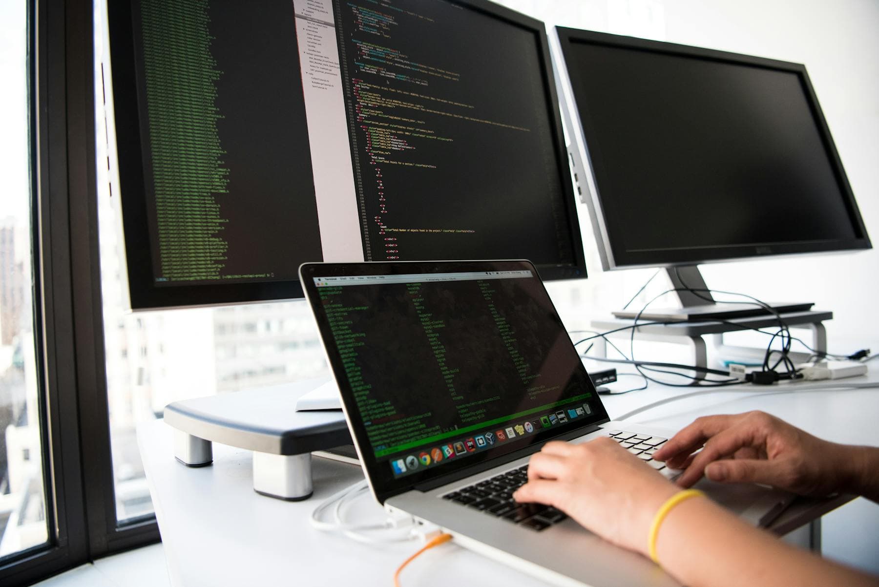 Developer writes code on laptop in front of multiple monitors